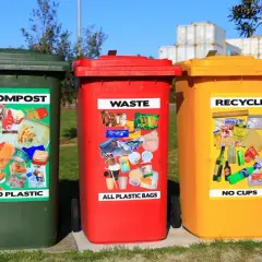 Trzy kosze na śmieci: zielony (kompost), czerwony (odpady) i żółty (recykling), z naklejkami pokazującymi przykładowe odpady.